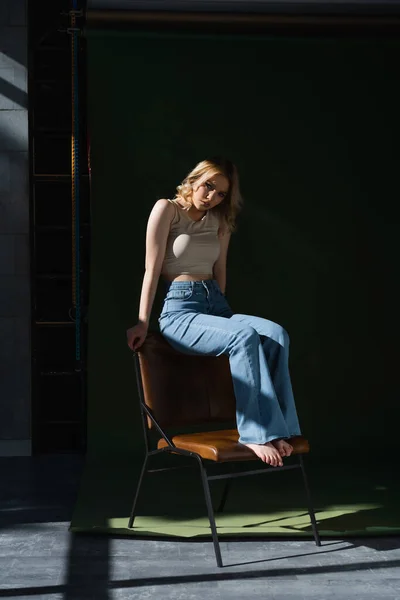 Full length view of young barefoot woman in jeans and top posing on chair on dark background — Stockfoto