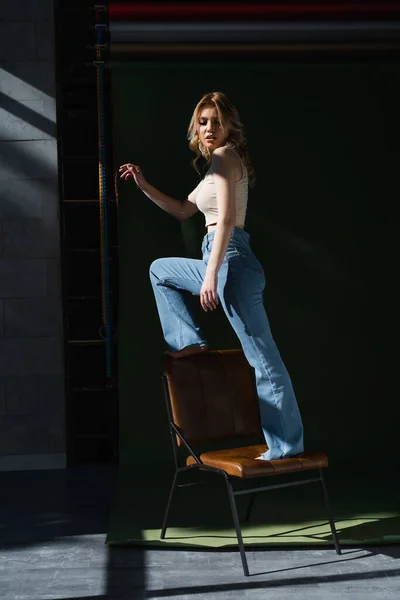 Vista completa de la mujer delgada en jeans de pie en silla sobre fondo oscuro - foto de stock