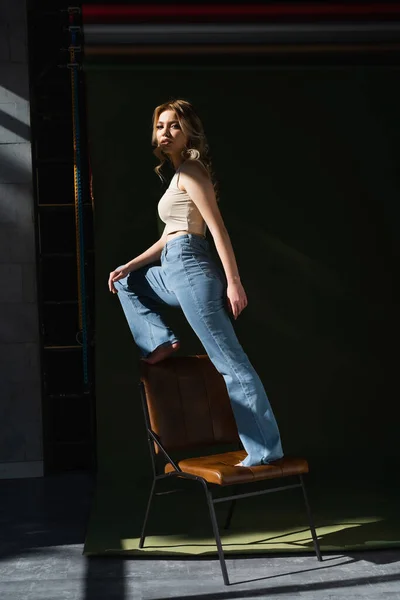 Pleine longueur de femme pieds nus en jeans regardant la caméra tout en se tenant sur la chaise sur fond sombre — Photo de stock