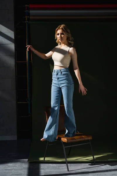 Full length of woman in beige top and jeans standing on chair in lighting on dark background — стоковое фото