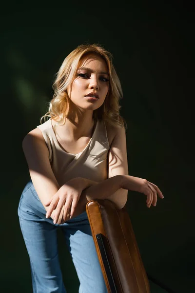 Jovem mulher no topo bege e jeans inclinados na cadeira e olhando para a câmera no fundo escuro — Fotografia de Stock