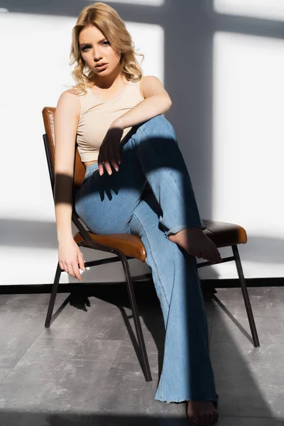 Full length view of slim woman in jeans sitting on chair near white wall with shadows — стоковое фото