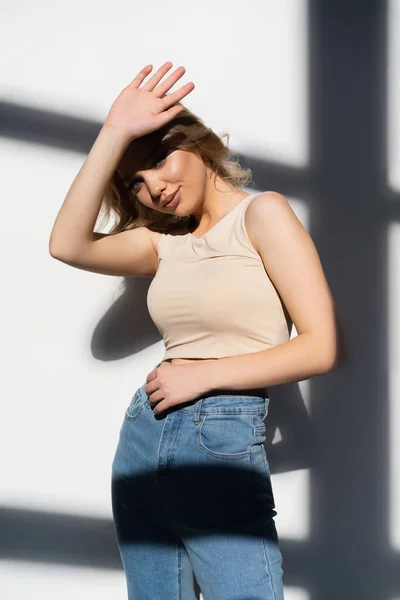Молода жінка в джинсах дивиться на камеру з-під руки на білому тлі з тінями — стокове фото