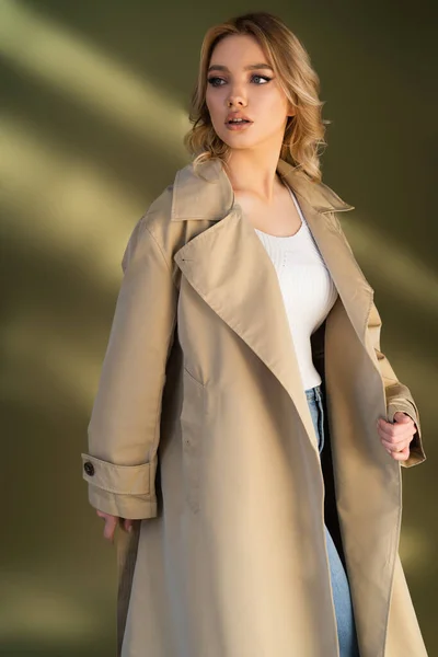 Femme aux cheveux ondulés regardant loin tout en posant en trench coat sur fond beige — Photo de stock