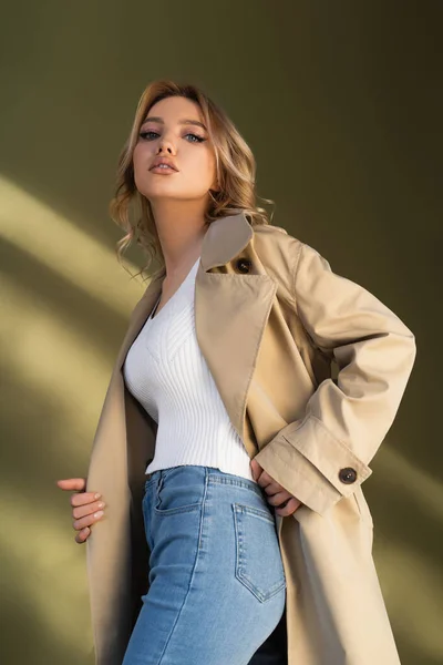 Low angle view of pretty woman in trench coat and jeans looking at camera on beige background — Photo de stock