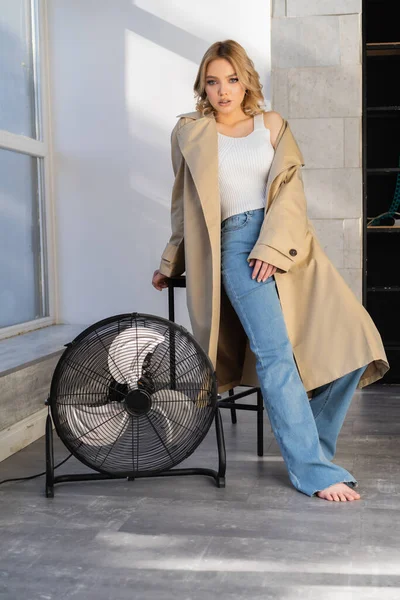 Ganzkörperansicht einer sinnlichen barfüßigen Frau in Jeans und Trenchcoat, die in der Nähe eines riesigen elektrischen Ventilators posiert — Stockfoto