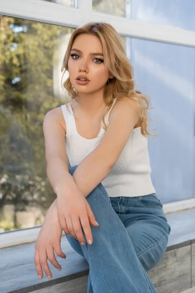 Young woman in jeans sitting near window and looking at camera — Stock Photo