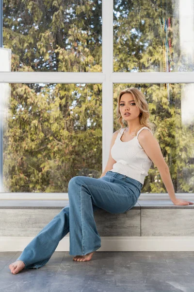 Full length view of slim barefoot woman in white tank top and jeans sitting on windowsill — Foto stock