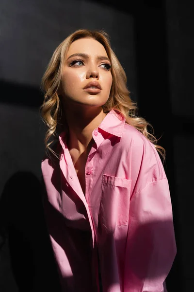 Junge Frau im rosafarbenen Hemd schaut weg, während sie im Licht auf schwarzem Hintergrund posiert — Stockfoto