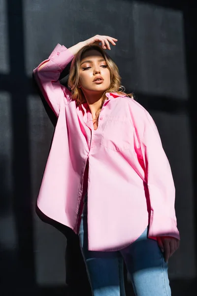 Pretty woman in pink shirt standing with hand above head near black wall with shadows — Foto stock