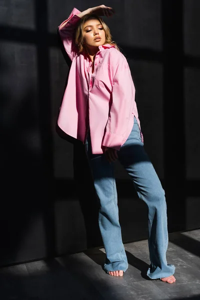 Vue pleine longueur de la femme à la mode pieds nus posant avec la main au-dessus de la tête près du mur noir avec des ombres — Photo de stock