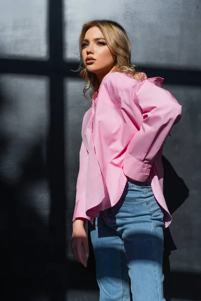 Stylish woman in pink shirt standing with hand in pocket of jeans near black wall with shadows — Photo de stock