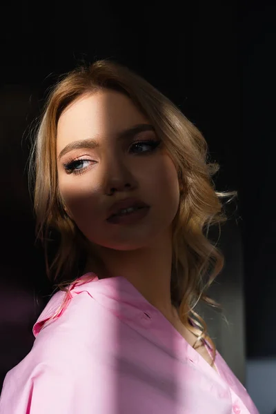 Portrait de charmante femme aux cheveux ondulés regardant loin dans la lumière sur fond sombre — Photo de stock