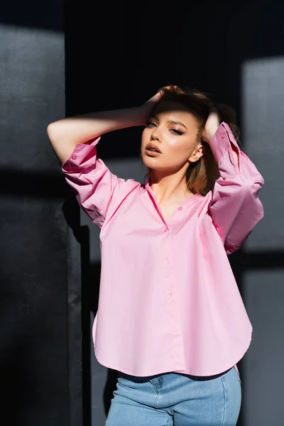 Charming woman in pink shirt posing with hands on head near black wall with shadows — стоковое фото