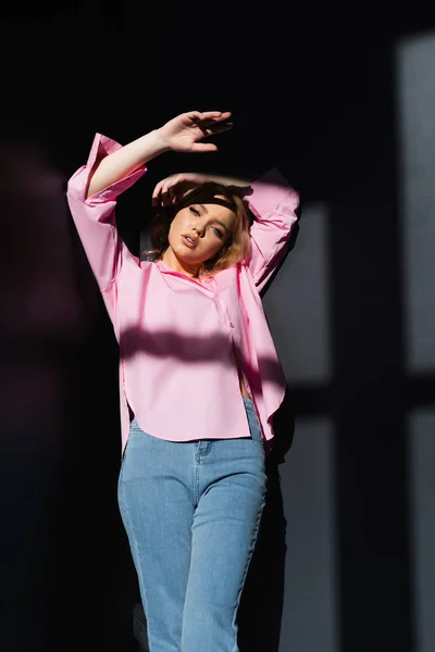 Young and stylish woman standing with hands above head on black background with shadows — Stockfoto