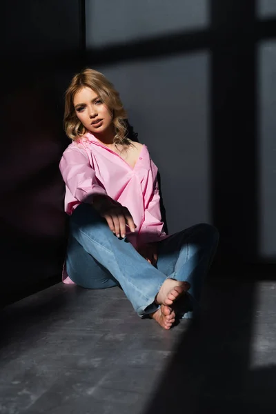 Полный вид босиком женщины в джинсах и розовой рубашке, сидящей в углу возле черных стен — стоковое фото
