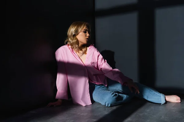 Full length view of barefoot woman in jeans sitting in lighting between black walls - foto de stock