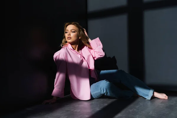 Full length of barefoot woman in pink shirt sitting on floor near black walls with shadows — Stockfoto