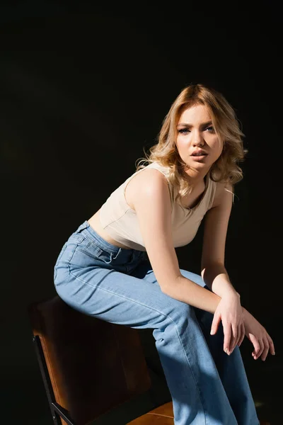 Young woman in jeans posing on chair and looking at camera on dark background — Fotografia de Stock