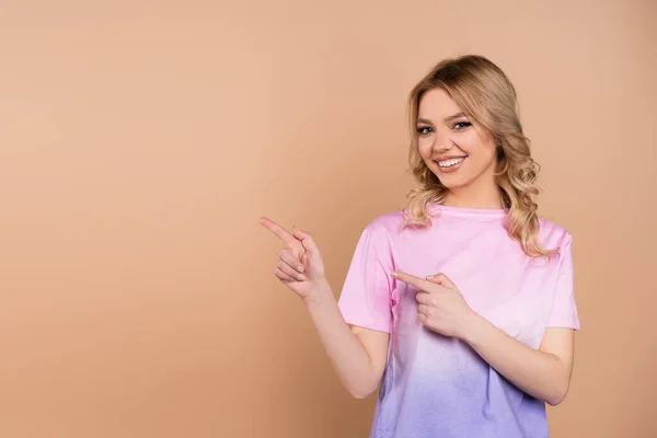 Cheerful woman pointing aside with fingers and looking at camera isolated on beige — Stock Photo