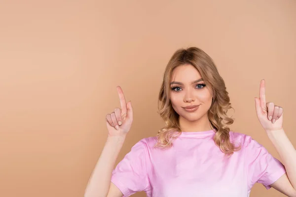 Smiling woman in t-shirt pointing up with fingers isolated on beige - foto de stock