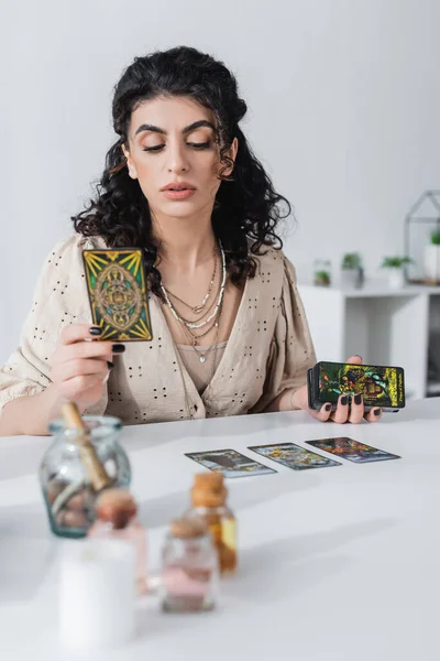 KYIV, UCRANIA - 23 de febrero de 2022: adivino gitano sosteniendo cartas del tarot cerca de suministros de brujería borrosa en casa - foto de stock