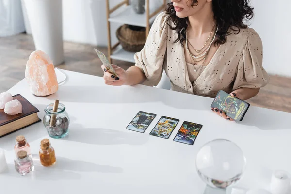 KYIV, UKRAINE - FEBRUARY 23, 2022: Cropped view of fortune teller holding tarot cards near book and crystals — Fotografia de Stock