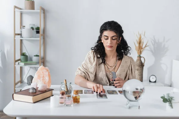 Adivino gitano sosteniendo cartas del tarot cerca de orbe borrosa en la mesa - foto de stock