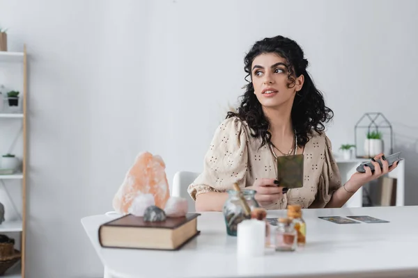 Sonriente gitano medio celebración del tarot tarjetas cerca de libro borroso y cristales en la mesa - foto de stock