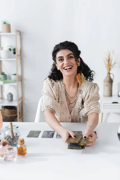 Sonriente adivino gitano mirando a la cámara y sosteniendo cartas del tarot en casa - foto de stock