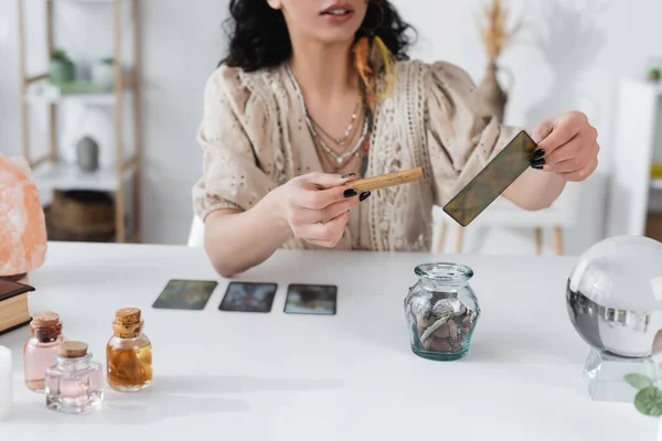 Vista recortada de adivino sosteniendo palo de salvia y tarjeta del tarot en casa - foto de stock