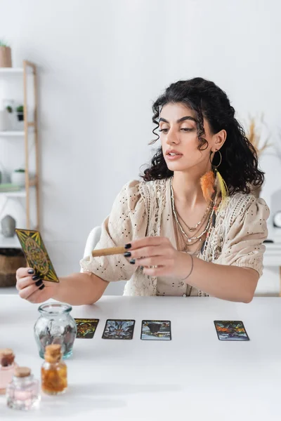 KYIV, UKRAINE - FEBRUARY 23, 2022: Young gypsy fortune teller holding tarot card and sage stick at home — Photo de stock