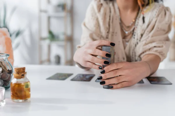 Cropped view of blurred medium holding tarot cards near jars on table — стоковое фото