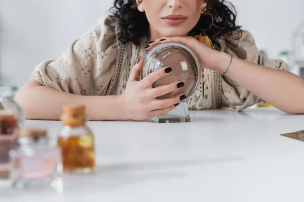 Cropped view of soothsayer touching orb near blurred witchcraft supplies on table — Stockfoto