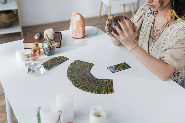 KYIV, UCRANIA - 23 de febrero de 2022: Vista recortada de orbe de celebración media cerca del tarot y velas en la mesa - foto de stock
