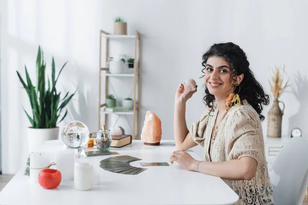 Positive gypsy soothsayer holding magic crystal and looking at camera near witchcraft supplies on table — Foto stock