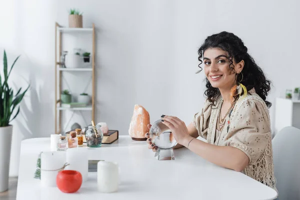 Sonriente adivino gitano mirando a la cámara cerca del orbe y velas en la mesa - foto de stock