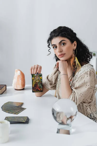 KYIV, UKRAINE - FEBRUARY 23, 2022: Gypsy fortune teller showing tarot card and looking at camera at home - foto de stock