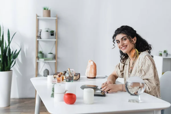 Diseuse de bonne aventure gitane souriante regardant caméra près de cartes de tarot, bougies et orbe à la maison — Photo de stock