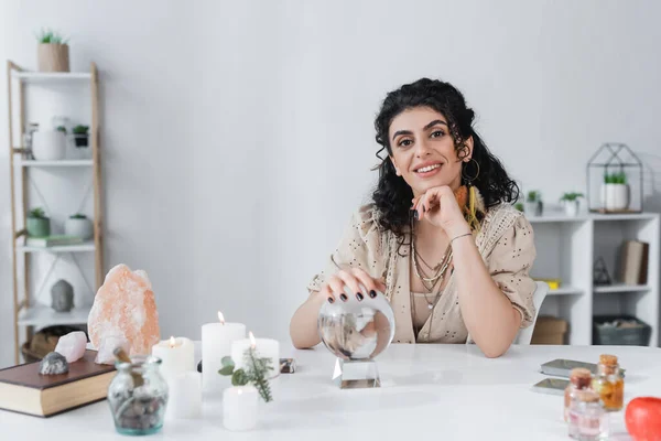 Medio gitano positivo mirando la cámara cerca del orbe, velas y cristales - foto de stock