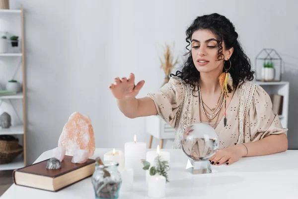 Adivino gitano sentado cerca de orbe mágico, velas y libro en casa - foto de stock
