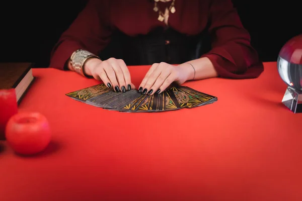 KYIV, UKRAINE - 23 FÉVRIER 2022 : Vue recadrée de diseur de bonne aventure tenant des cartes de tarot près de bougies et livre isolé sur noir — Photo de stock