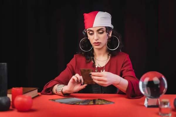 Adivino gitano mirando borrosa carta del tarot cerca de orbe de cristal en la mesa aislado en negro - foto de stock