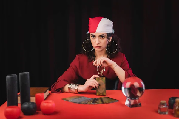 Gypsy fortune teller holding tarot and looking at camera near witchcraft supplies isolated on black — Stock Photo