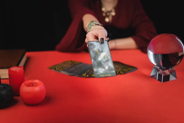 Ausgeschnittene Ansicht des verschwommenen Mediums mit Tarotkarte in der Nähe von Kerzen und Kugel isoliert auf schwarz — Stockfoto