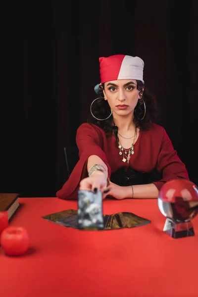 Gypsy fortune teller holding blurred tarot and looking at camera isolated on black — Stock Photo