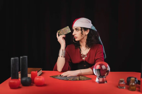 KYIV, UKRAINE - FEBRUARY 23, 2022: Young gypsy fortune teller holding tarot near witchcraft supplies on table isolated on black — Photo de stock