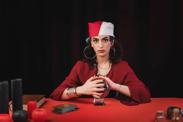 Gypsy fortune teller touching magic orb near tarot and candles isolated on black - foto de stock