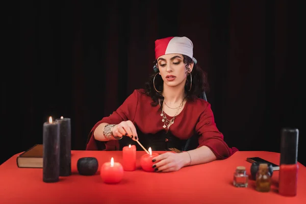 Gypsy soothsayer burning candles near book and witchcraft supplies on table isolated on black — Stockfoto