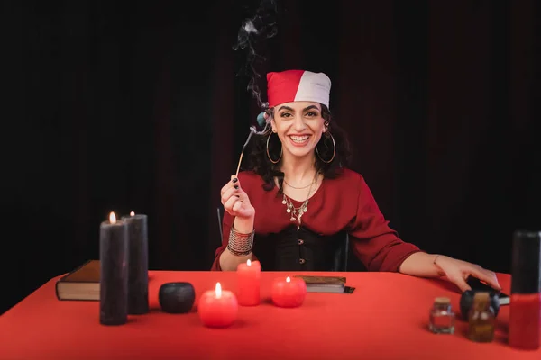 Cheerful gypsy soothsayer holding match near candles and blurred witchcraft supplies isolated on black - foto de stock